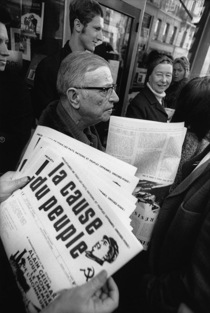 Le philosophe Jean-Paul Sartre avec Simone de Beauvoir (au fond, le regardant en souriant) distribuant le journal maoïste "La Cause du Peuple", interdit par le gouvernement français, à Paris en 1970. (© Bruno Barbey/Magnum Photos)